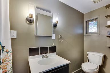Compact gray bathroom with white vanity and sink, mirrored medicine cabinet flanked by glass wall sconces, white toilet, small stained-glass window and floral shower curtain