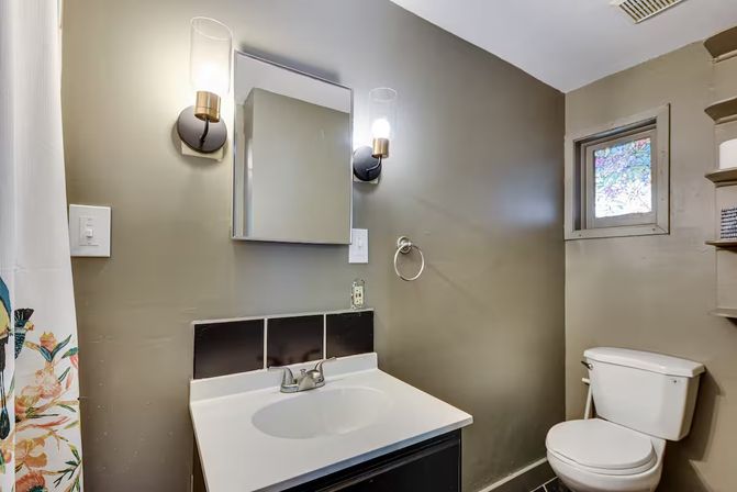 Compact gray bathroom with white vanity and sink, mirrored medicine cabinet flanked by glass wall sconces, white toilet, small stained-glass window and floral shower curtain