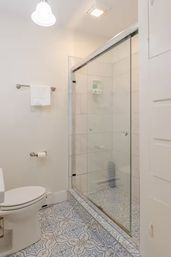 Bright small white bathroom with glass sliding walk-in shower, geometric blue-gray patterned tile floor, white toilet and wall-mounted towel rack