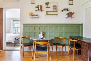 Sunlit dining nook with round gray table and four mid-century wooden chairs, green paneled wainscoting, floating shelves with books and plants, hardwood floor and a bedroom visible through a doorway.