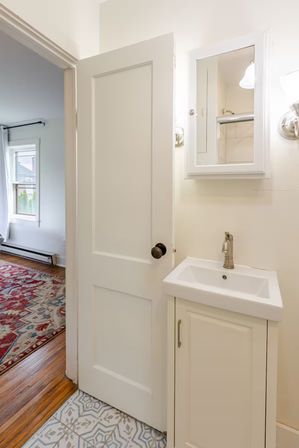 Cozy small white bathroom with compact vanity sink, brushed-nickel faucet, mirrored medicine cabinet and sconce, gray patterned floor tile; open door reveals hardwood bedroom with red area rug and window.