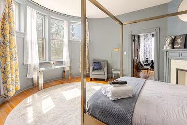 Sunlit Victorian-style bedroom with a gold four-poster bed topped with folded towels, gray walls, curved bay windows with yellow floral curtains, hardwood floors, round area rug and a cozy armchair