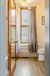 Cozy small bathroom with wood‑frame window and natural light, gray curtain, white toilet, glass‑paneled wooden door and wood‑look vinyl plank flooring.
