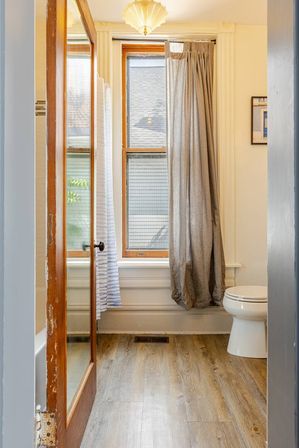 Cozy small bathroom with wood‑frame window and natural light, gray curtain, white toilet, glass‑paneled wooden door and wood‑look vinyl plank flooring.