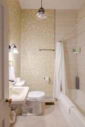 Cozy residential bathroom interior with yellow floral wallpaper, white pedestal sink and toilet, bathtub with white shower curtain, brass fixtures, glass pendant light and beige tiled floor.