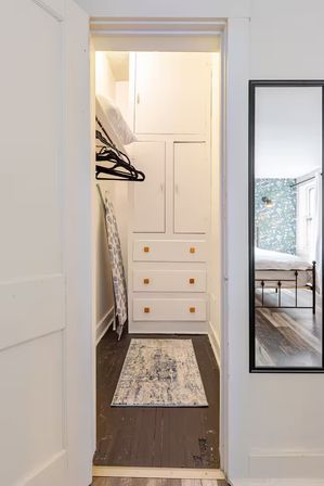 Cozy narrow walk-in closet with white built-in cabinets and drawers, black hangers on a rod, patterned ironing board leaning against the wall, and a small distressed dark-wood floor rug; full-length mirror reflects an adjacent bedroom with floral wallpaper and a metal bed.