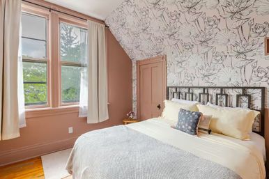 Sunlit Victorian-style bedroom with black-and-white floral wallpaper, blush-pink walls and trim, large double window with cream curtains, iron headboard, cream bedding and patterned throw pillows.