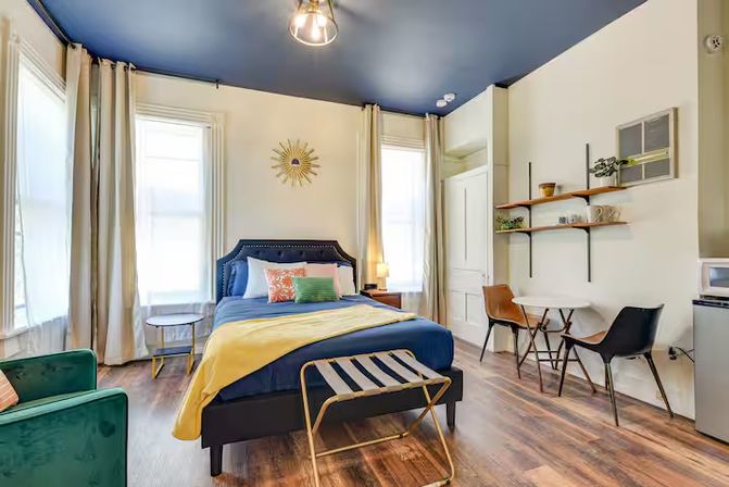 Bright modern studio bedroom in an urban apartment with a navy ceiling, queen bed dressed in blue linens and a yellow throw, sunburst wall art, tall sheer-curtained windows, small dining table with chairs, open shelving, green accent chair, and warm wood floors.