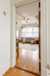 Bright cozy living room seen through a doorway with hardwood floors, tufted gray sofa, two yellow ottomans, sheer-curtained large windows, patterned area rug and ceiling fan.