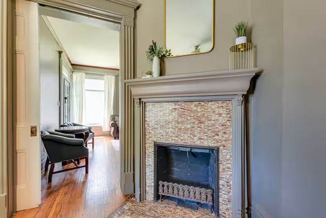 Cozy sunlit living room with decorative tiled fireplace and mantel mirror, potted plants, hardwood floors, and an adjacent sitting area with armchairs.