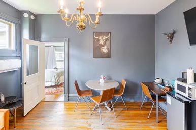 Cozy urban apartment dining nook with gray walls, brass chandelier, round marble-top table and four tan mid-century chairs on wooden floors, small kitchenette with microwave and mini fridge, bull artwork on the wall and open door to a bedroom.