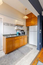 Cozy urban studio kitchenette with warm oak cabinets, granite countertop and double sink, turquoise coffee maker and microwave, open wood shelves with glassware, white refrigerator, mosaic tile backsplash, pendant light and gray runner rug.