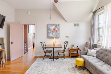 Sunlit cozy living room with hardwood floors, patterned area rug, gray tufted sofa and bright yellow ottoman, small wooden dining table with two black metal chairs under floral wall art, sheer curtains over a large window.