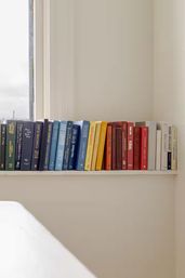 Slim white shelf beneath a window holding a rainbow of hardcover books arranged by color against a neutral beige bedroom wall