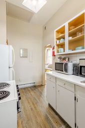 Compact bright galley apartment kitchen with white cabinets and open shelves holding pastel dishes, countertop microwave and coffee maker, electric coil stove, wood-look vinyl floor and arched doorway to living room.