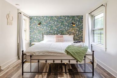 Cozy bright bedroom with black iron bedframe, crisp white linens, floral green accent wallpaper, wall-mounted lamps, two windows, and a folded green knit throw at the foot of the bed.