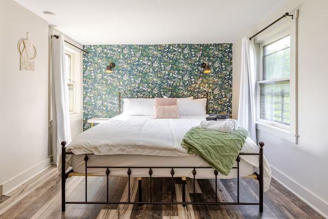 Cozy bright bedroom with black iron bedframe, crisp white linens, floral green accent wallpaper, wall-mounted lamps, two windows, and a folded green knit throw at the foot of the bed.