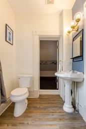 Cozy residential bathroom with white toilet and pedestal sink, brass globe vanity lights, blue accent wall with mirror, wood-look plank flooring, and an open doorway to a small walk-in closet with hangers.