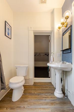 Cozy residential bathroom with white toilet and pedestal sink, brass globe vanity lights, blue accent wall with mirror, wood-look plank flooring, and an open doorway to a small walk-in closet with hangers.