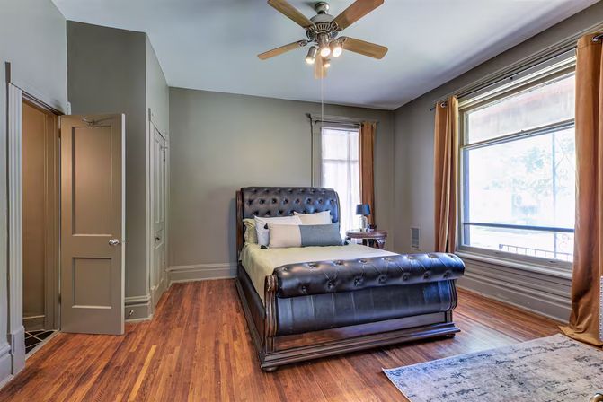Bright, cozy bedroom with a tufted dark leather sleigh bed, hardwood floors, ceiling fan, large window with brown curtains and a bedside table.