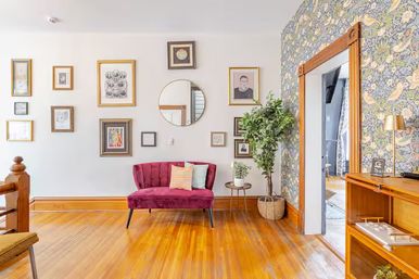 Bright entryway sitting area with pink velvet loveseat, gallery wall of framed art and round mirror, floral wallpaper accent wall, tall potted plant, and polished hardwood floors.