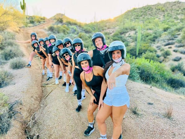 Smiling group of women wearing helmets and pink bandanas lined up on a Sonoran Desert trail with saguaro cacti; front woman shows a ring.