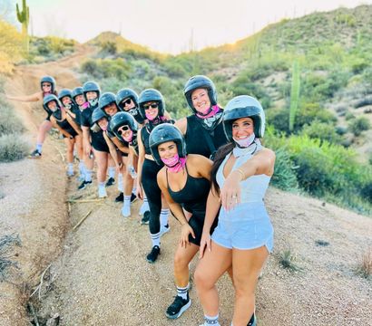 Smiling group of women wearing helmets and pink bandanas lined up on a Sonoran Desert trail with saguaro cacti; front woman shows a ring.