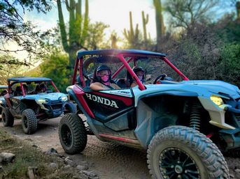 Two off-road UTV side-by-sides with masked riders on a dusty Sonoran Desert trail at sunset, saguaro cacti and rocky terrain in the background