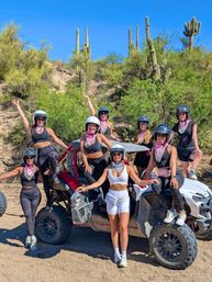 Group of eight women in helmets and bandanas posing on and around a red-and-white off-road UTV amid saguaro cacti and desert scrub in the Sonoran Desert under a bright blue sky