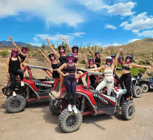 Group of women in helmets and pink bandanas cheering and posing on red off-road UTVs in a desert landscape with saguaro cacti, dirt trail and bright blue sky.