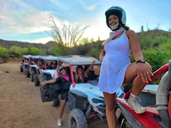 Smiling woman in a helmet poses on a red UTV while a group lines up in blue off-road buggies along a cactus-studded desert trail