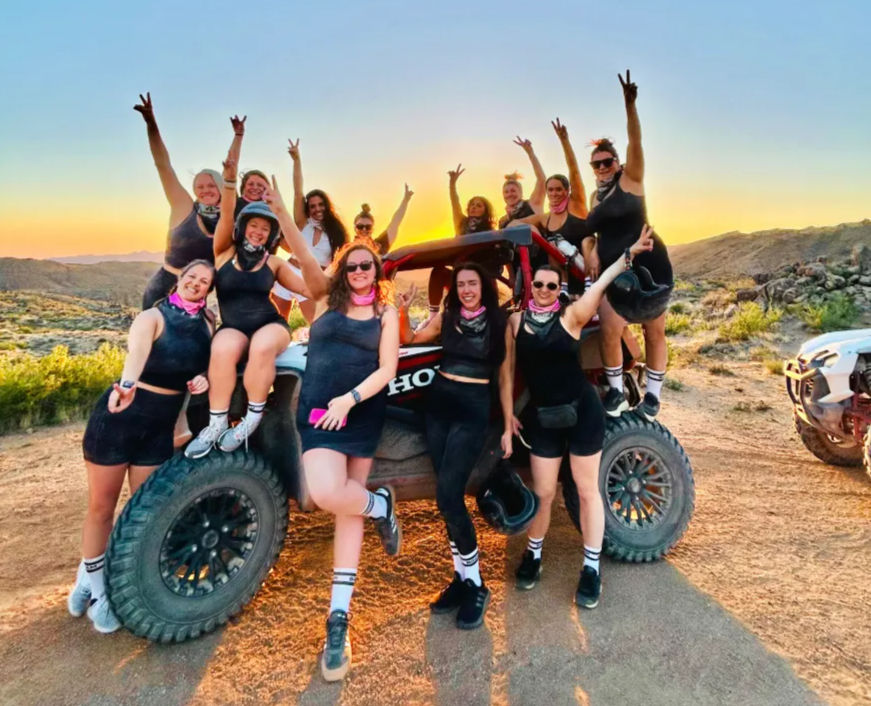 Group of women posing on and around an off-road UTV at sunset in a rocky desert landscape, cheering and flashing peace signs.