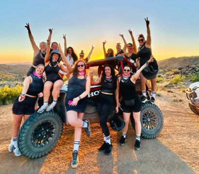 Group of women posing on and around an off-road UTV at sunset in a rocky desert landscape, cheering and flashing peace signs.