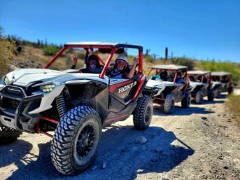 Line of white-and-red side-by-side off-road UTVs on a sunny desert trail with helmeted drivers and goggles, one waving, with cacti and scrubby hills in the background.