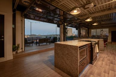 Industrial open-plan loft kitchen with large granite island, stainless appliances, wood floors and pendant lights opening to a rooftop patio with string lights and cushioned outdoor seating overlooking a city skyline at dusk.