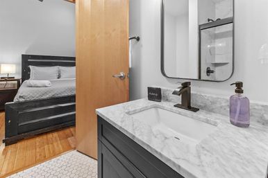 Modern bathroom with marble vanity and undermount sink, oil-rubbed bronze faucet, rectangular mirror and purple soap dispenser; hexagon tile floor and partially open wooden door revealing a cozy bedroom with platform bed, white linens and hardwood floors.