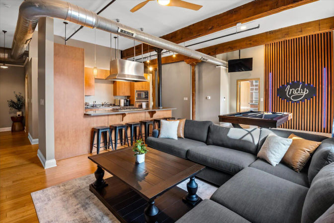 Industrial-style open-concept loft with gray sectional, dark wood coffee table, kitchen bar with metal stools, exposed ductwork and beams, pool table and “Indy” neon sign.