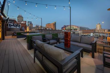 Downtown rooftop patio at dusk with cushioned outdoor sofas, string lights, wood tables and a skyline view including an illuminated arena/stadium.