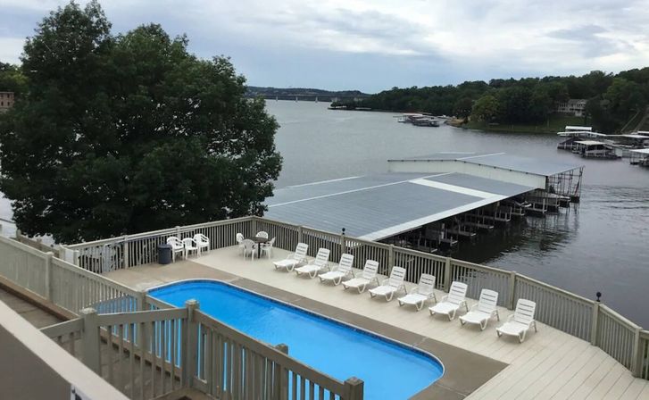 Lakefront pool deck with bright blue rectangular pool, row of white lounge chairs and patio table, overlooking covered boat docks and calm lake under a cloudy sky