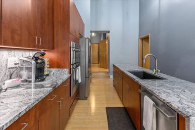 Contemporary galley kitchen with gray veined stone countertops, warm wood cabinets, stainless steel appliances, single-basin undermount sink on a long island, coffee maker on the counter, and light hardwood floors.