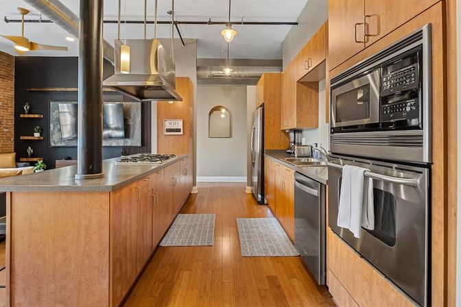 Open-concept urban loft kitchen with warm wood cabinets, stainless steel appliances, central island with gas cooktop and hood, exposed ductwork and hardwood floors.