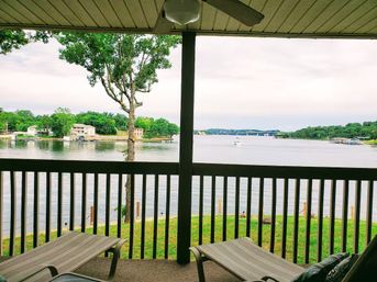 Relaxing lakefront covered porch with lounge chairs and railing, overlooking a calm lake with a boat in the distance and tree-lined shoreline with waterfront homes