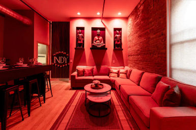 Red-lit modern lounge with large sectional sofa, exposed brick wall, recessed lights, Buddha and warrior statues in wall niches, nested round coffee tables, bar seating and hardwood floor.