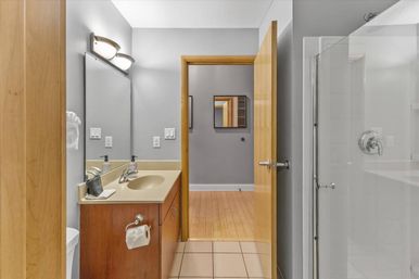 Bright, tidy modern residential bathroom with wood vanity and beige countertop sink, wall mirror with sconce lights, glass shower door, light gray walls and tiled floor, open wood door showing hardwood hallway.