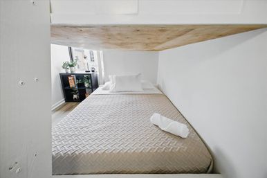 Cozy minimalist urban studio sleeping nook with full bed, beige quilt, white pillow and rolled towel under a wooden loft, small shelf with plants by the window.