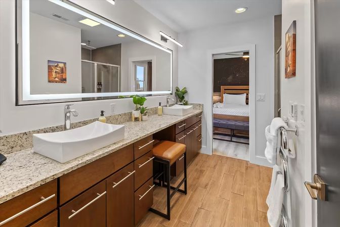 Sleek modern bathroom with long granite countertop, two white vessel sinks, wood cabinetry, large backlit mirror, leather vanity stool, potted plants, towel rack and doorway leading to a bedroom.