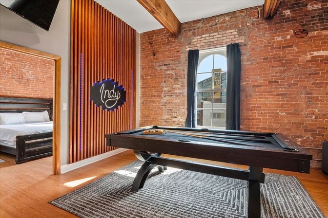 Urban industrial loft game room with a sleek black pool table on a patterned rug, exposed red brick walls, wooden beams, arched window with city view, slatted wood accent wall with neon sign, and an adjacent bedroom.
