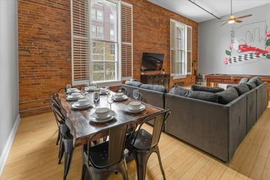 Stylish urban loft living and dining area with exposed brick walls, tall arched windows with shutters, hardwood floors, a rustic wood dining table set for six with white dishes and metal chairs, a large gray sectional sofa facing a TV, and a colorful wall mural above a pool table.