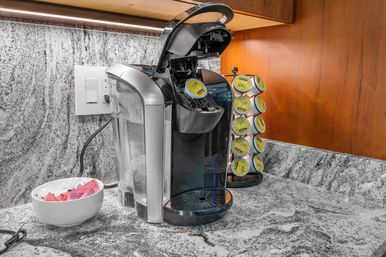 Single-serve coffee maker with open lid on a marble kitchen countertop, beside a rotating rack of yellow coffee pods and a white bowl of sugar and sweetener packets under warm under-cabinet lighting.