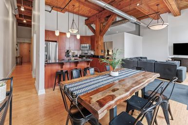 Industrial-chic open-concept loft apartment with exposed wooden beams and ductwork, hardwood floors, live-edge dining table with patterned runner, black metal chairs, kitchen island with bar stools and stainless-steel appliances, pendant lighting and a gray sectional sofa.
